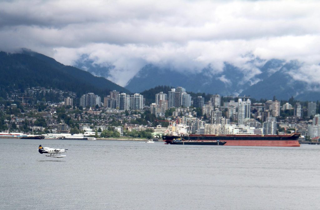 Seaplane flying over Nanaimo, BC waterfront with city skyline and mountains in the background.